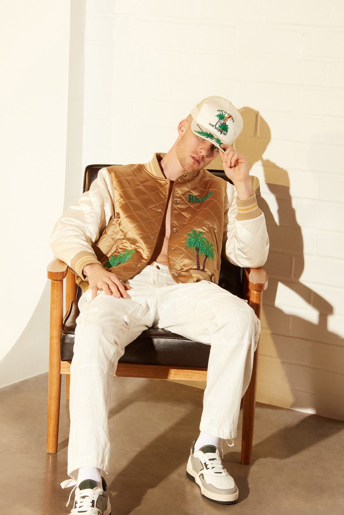 A male model sitting in a chair in a lazy style wearing Rhude jerkin and a Rhude cap