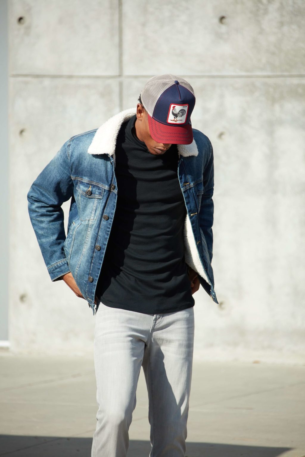 A male model with trousers , tshirt and a short jean over coat with a cap